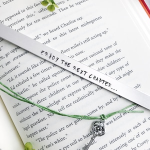 Op de afbeelding: Een zilveren bladwijzer met een groen touwtje en een zilveren sleutelbedel. De bladwijzer heeft de tekst "Enjoy the next chapter..." erop staan. De bladwijzer ligt op een open boek.