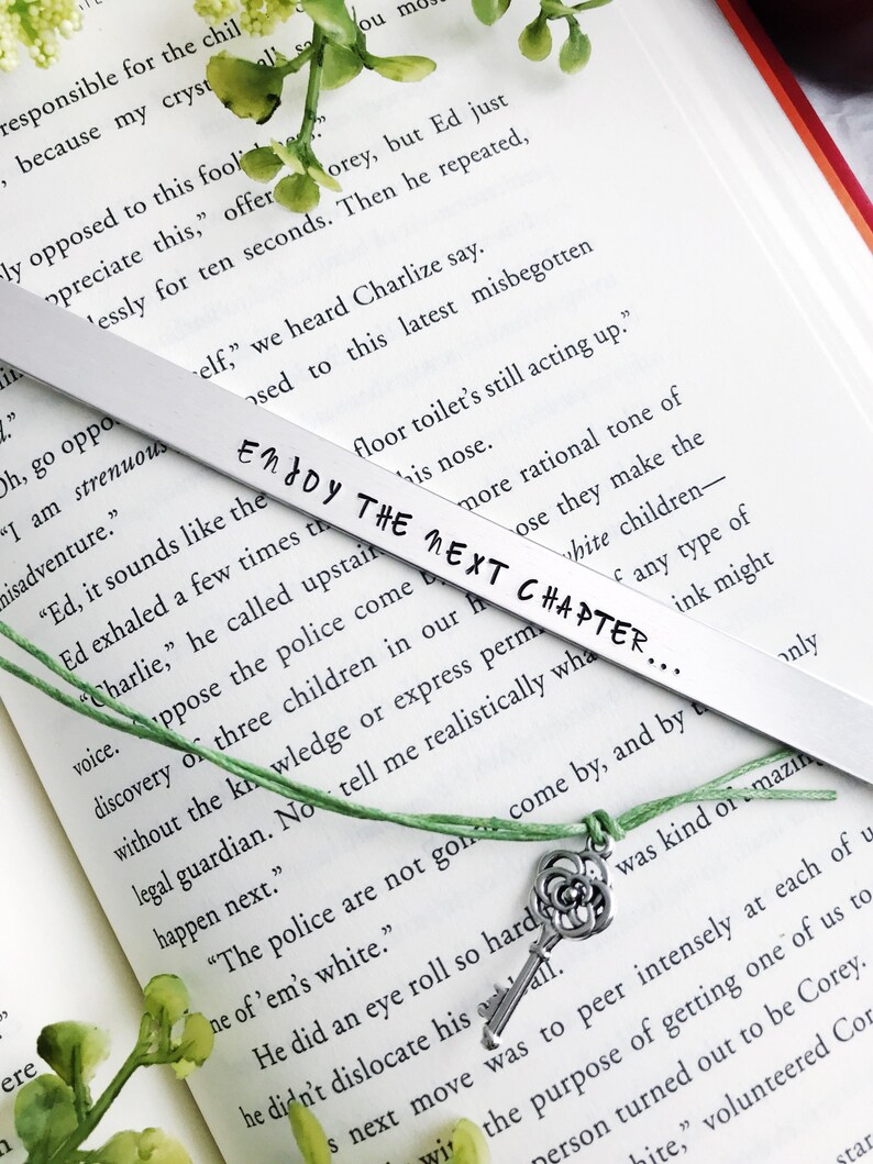 Op de afbeelding: Een zilveren bladwijzer met een groen touwtje en een zilveren sleutelbedel. De bladwijzer heeft de tekst "Enjoy the next chapter..." erop staan. De bladwijzer ligt op een open boek.