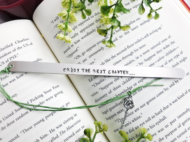 Op de afbeelding: Een zilveren metalen bladwijzer met de tekst "Enddy The Next Chapter..." erop gegraveerd. De bladwijzer is bevestigd aan een groen touwtje met een zilveren sleutelbedel.