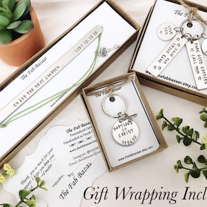 Op de afbeelding: Drie cadeaudozen met gepersonaliseerde metalen bladwijzers en sleutelhangers. De dozen zijn bruin met witte tekst die "The Fab Bazaar" luidt. De bladwijzers en sleutelhangers bevatten verschillende inspirerende berichten en data. De dozen staan op een wit oppervlak met een kleine groene plant op de achtergrond.