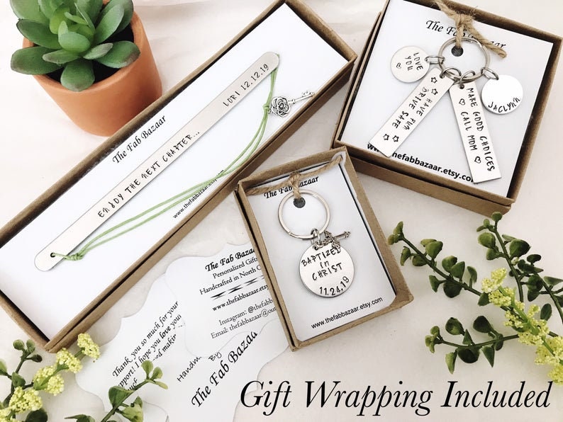 Op de afbeelding: Drie cadeaudozen met gepersonaliseerde metalen bladwijzers en sleutelhangers. De dozen zijn bruin met witte tekst die "The Fab Bazaar" luidt. De bladwijzers en sleutelhangers bevatten verschillende inspirerende berichten en data. De dozen staan op een wit oppervlak met een kleine groene plant op de achtergrond.