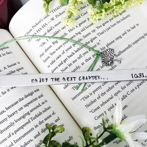 Op de afbeelding: Een zilveren bladwijzer met de tekst "Enjoy the next chapter..." en de datum "10.31.18" erop gegraveerd. De bladwijzer ligt op een boek met een groene plant en een zilveren boomhanger.