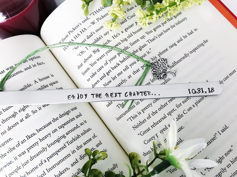Op de afbeelding: Een zilveren bladwijzer met de tekst "Enjoy the next chapter..." en de datum "10.31.18" erop gegraveerd. De bladwijzer ligt op een boek met een groene plant en een zilveren boomhanger.