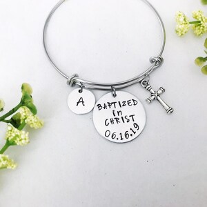 Puede incluir: Pulsera de brazalete de plata con tres dijes: un dije redondo con la letra "A", un dije redondo con el texto "BAPTIZED in CHRIST 06.16.19" y un peque&ntilde;o dije de cruz.
