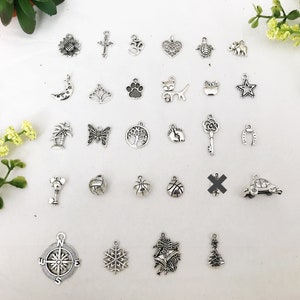 Op de afbeelding: Een verzameling van 27 zilveren bedelarmbanden met verschillende ontwerpen, waaronder een zonnebloem, een kruis, een om-symbool, een hart, een schildpad, een olifant, een halve maan, een lotusbloem, een pootprint, een kat, een appel, een ster, een palmboom, een vlinder, een levensboom, een voetafdruk, een sleutel, een hoefijzer, een auto, een basketbal, een kompas, een sneeuwvlok, een bel en een kerstboom.
