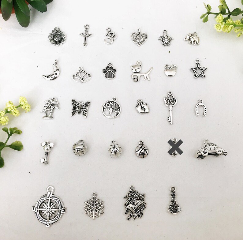 Op de afbeelding: Een verzameling van 27 zilveren bedelarmbanden met verschillende ontwerpen, waaronder een zonnebloem, een kruis, een om-symbool, een hart, een schildpad, een olifant, een halve maan, een lotusbloem, een pootprint, een kat, een appel, een ster, een palmboom, een vlinder, een levensboom, een voetafdruk, een sleutel, een hoefijzer, een auto, een basketbal, een kompas, een sneeuwvlok, een bel en een kerstboom.