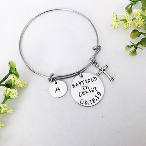 Puede incluir: Un brazalete de plata con un dije que dice "Bautizado en Cristo 06.16.19" y un dije con la letra "A". El brazalete tambi&eacute;n tiene un peque&ntilde;o dije de cruz de plata.