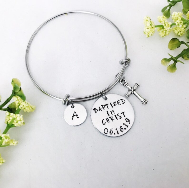 Puede incluir: Un brazalete de plata con un dije que dice "Bautizado en Cristo 06.16.19" y un dije con la letra "A". El brazalete tambi&eacute;n tiene un peque&ntilde;o dije de cruz de plata.