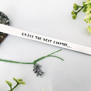 Op de afbeelding: Zilveren bladwijzer met de tekst "Enjoy the next chapter..." gegraveerd en bevestigd aan een groen touwtje met een palmboombedel.