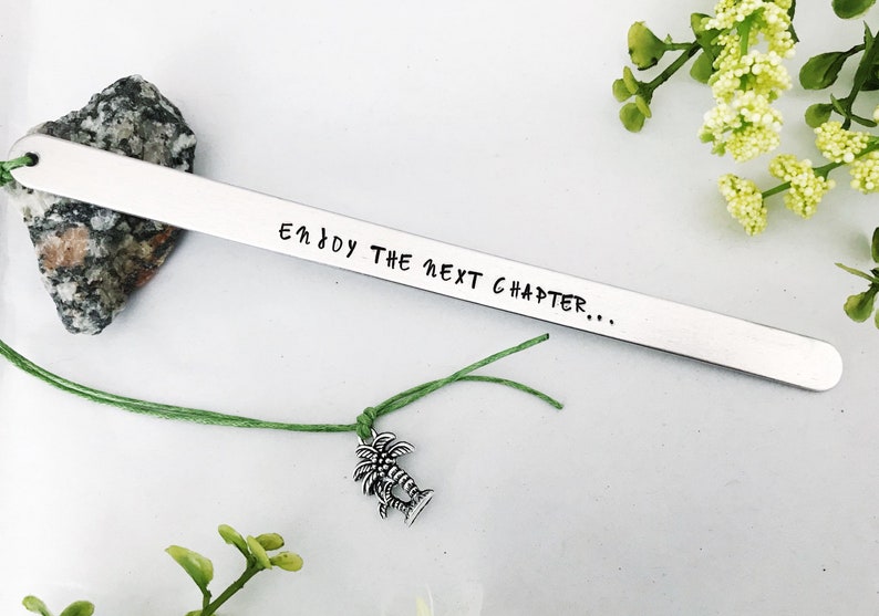 Op de afbeelding: Zilveren bladwijzer met de tekst "Enjoy the next chapter..." gegraveerd en bevestigd aan een groen touwtje met een palmboombedel.