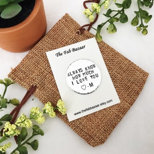 May include: Silver round charm with the text "Always know how much I love you -M" on a hessian background. The charm is in a small hessian bag with a brown drawstring.
