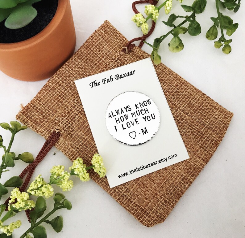 May include: Silver round charm with the text "Always know how much I love you -M" on a hessian background. The charm is in a small hessian bag with a brown drawstring.