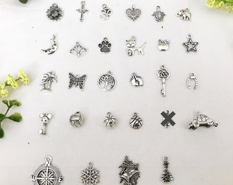 Puede incluir: Una colección de 27 colgantes de plata con diferentes diseños, incluyendo un girasol, una cruz, un símbolo om, un corazón, una tortuga, un elefante, una luna creciente, una flor de loto, una huella de pata, un gato, una manzana, una estrella, una palmera, una mariposa, un árbol de la vida, una huella de pie, una llave, una herradura, un coche, un balón de baloncesto, una brújula, un copo de nieve, una campana y un árbol de Navidad.