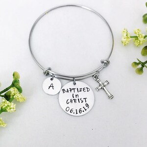 Puede incluir: Un brazalete de plata con tres dijes: un dije redondo con el texto "BAPTIZED in CHRIST 06.16.19", un dije redondo con la letra "A" y un dije de cruz de plata.