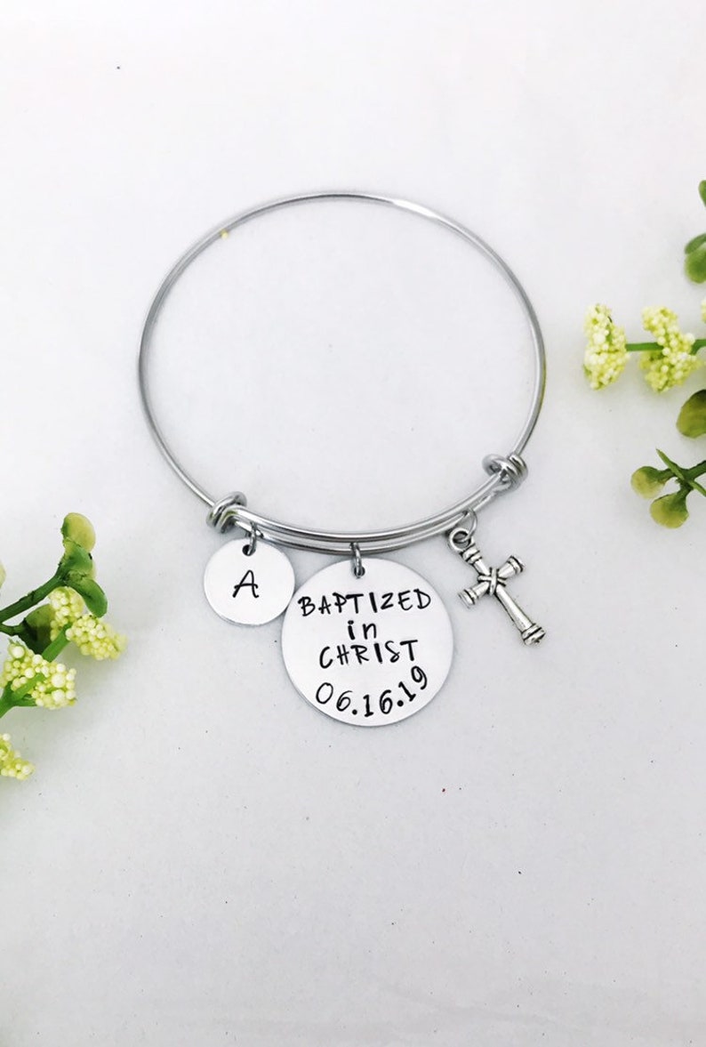 Puede incluir: Un brazalete de plata con tres dijes: un dije redondo con el texto "BAPTIZED in CHRIST 06.16.19", un dije redondo con la letra "A" y un dije de cruz de plata.