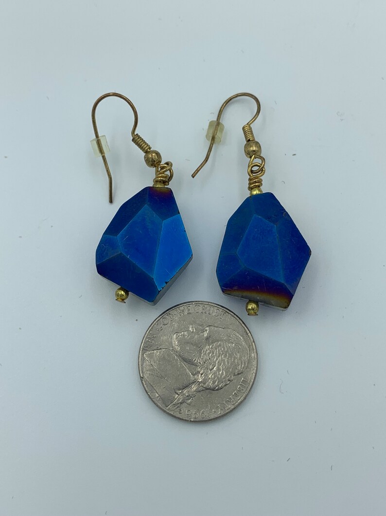 Peut inclure: Une paire de boucles d'oreilles en pierre bleue avec du fil d'or enroul&eacute; autour du sommet de la pierre et des crochets dor&eacute;s. Les boucles d'oreilles sont &agrave; peu pr&egrave;s de la taille d'une pi&egrave;ce de 25 cents am&eacute;ricaine.