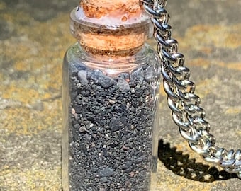 Puede incluir: Una pequeña botella de vidrio llena de arena negra, con un tapón de corcho y una cadena plateada unida. La botella cuelga de la cadena.