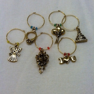 May include: Set of six gold-tone wine glass charms with different decorative charms, including an angel, a ring, a bouquet of flowers, a heart with "I love you" written on it, a balloon, and a cake.