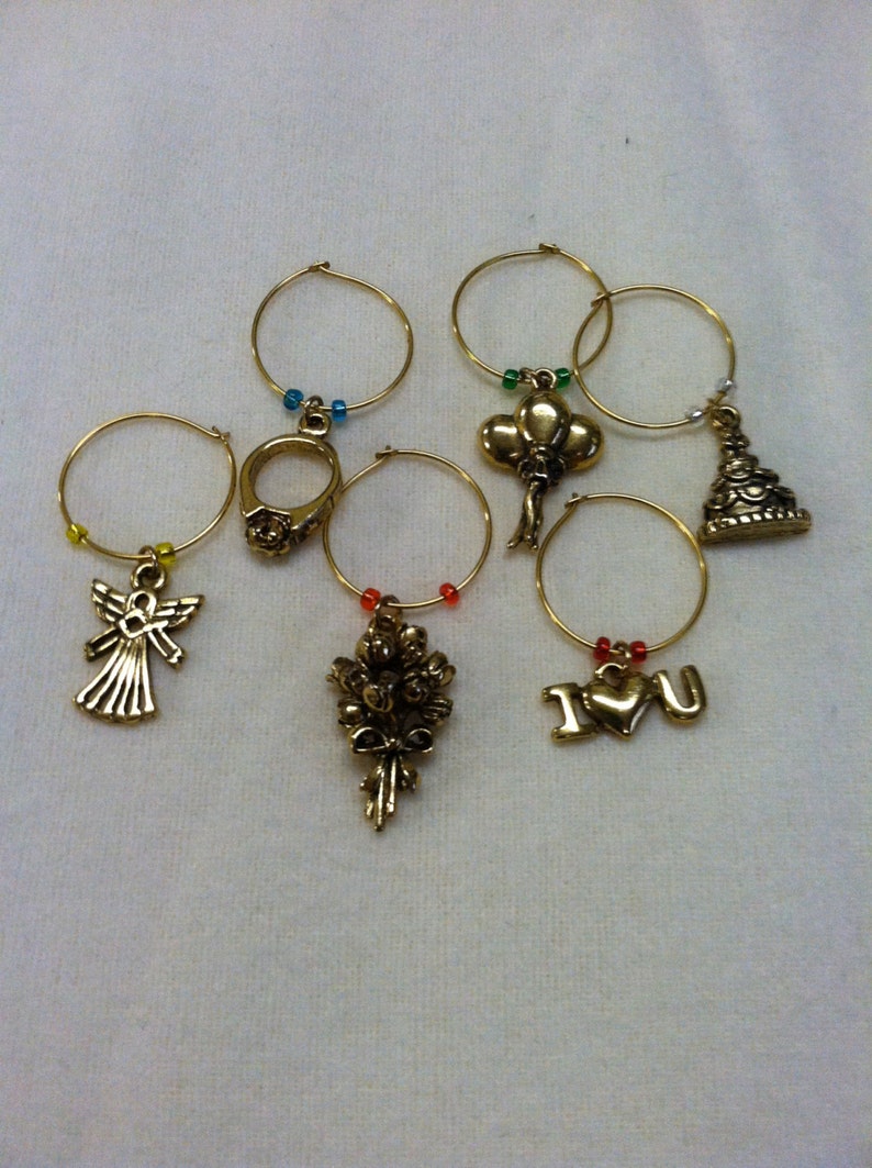 May include: Set of six gold-tone wine glass charms with different decorative charms, including an angel, a ring, a bouquet of flowers, a heart with "I love you" written on it, a balloon, and a cake.