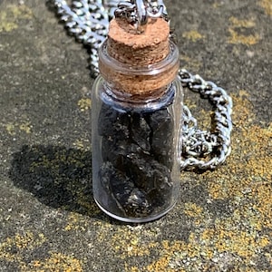 Op de afbeelding: Een kleine glazen flesjes hanger ketting gevuld met donkere, ruwe stenen. De fles heeft een kurken stopper en hangt aan een zilverkleurige ketting. De ketting wordt gepresenteerd op een getextureerd, grijs en geel oppervlak.
