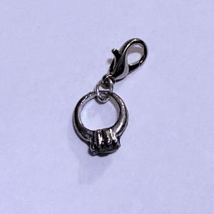 May include: A silver-coloured charm in the shape of a ring with a small gemstone. The charm is attached to a lobster clasp, allowing it to be easily added to a bracelet or necklace. The ring charm is a jewellery accessory.