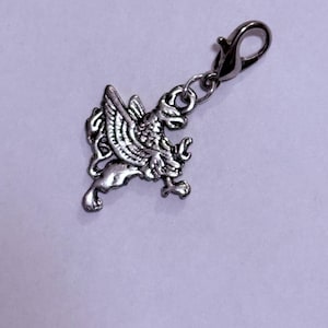 May include: A silver griffin charm, a mythical beast with a lion's body and an eagle's head and wings. The charm is attached to a lobster clasp, ideal for a bracelet or necklace. The charm is made of a silver-colored metal.