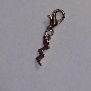 May include: A silver-colored charm with a lightning bolt design. The charm is attached to a lobster clasp with a small chain. The lightning bolt is a zig-zag shape. The charm is likely made of metal.