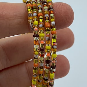 Könnte beinhalten: Mehrfarbiges Perlenarmband mit gelben, orangen, braunen und weißen Perlen. Das Armband besteht aus kleinen, runden Perlen und ist um einen Finger gewickelt.