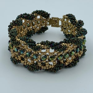 Op de afbeelding: Een gouden en groene kralen armband met een sluiting. De armband is gemaakt met kleine, ronde kralen en heeft een geometrisch patroon.