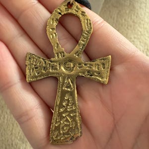 May include: A gold-colored Ankh pendant with intricate details and a black cord necklace.