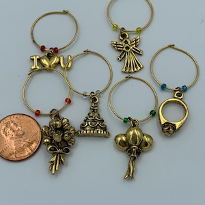May include: Set of six gold-tone wine glass charms with various designs, including an angel, a ring, a cake, balloons, a bouquet of flowers, and a charm with the words "I love you".