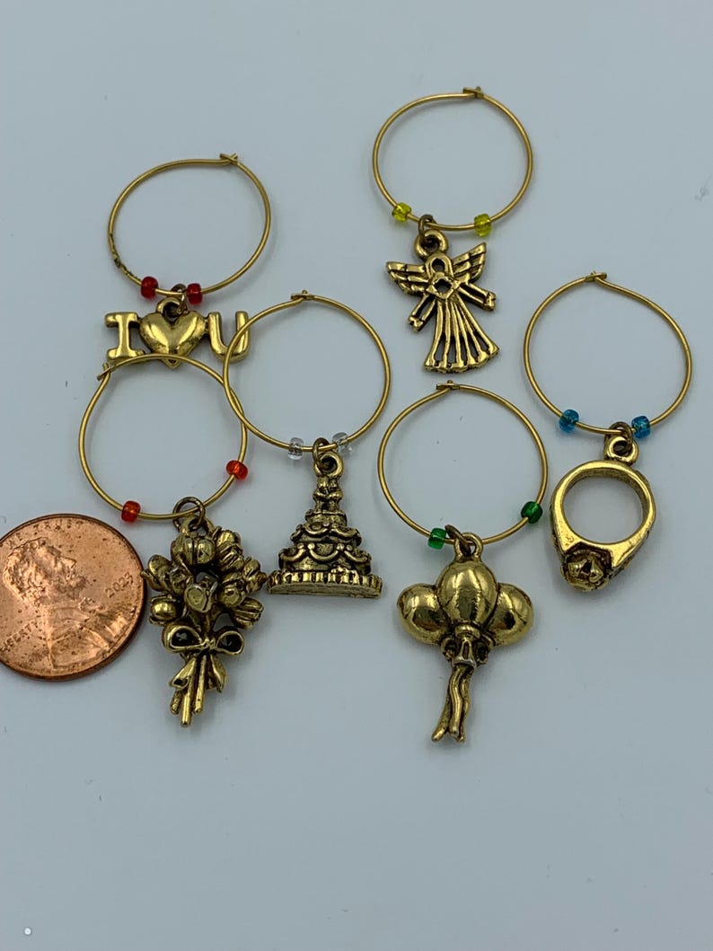 May include: Set of six gold-tone wine glass charms with various designs, including an angel, a ring, a cake, balloons, a bouquet of flowers, and a charm with the words "I love you".