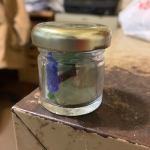 Op de afbeelding: Een klein glazen potje met een gouden deksel gevuld met kleurrijk zeeglas. Het potje staat op een houten oppervlak.