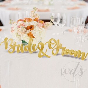 May include: Gold "Bride & Groom" sign with a white and peach floral arrangement on a white tablecloth.