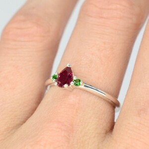 May include: A silver ring with a pear-shaped red gemstone and two smaller green gemstones.