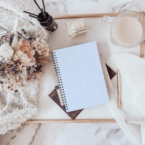 May include: A light blue spiral notebook with a white dotted pattern rests on a tray with a gold pen, a glass of liquid, and a bouquet of flowers. A small perfume bottle and a reed diffuser are also on the tray.