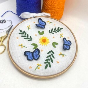 May include: Embroidered hoop art featuring three blue butterflies, a yellow sunflower, and green leaves on a white linen background.