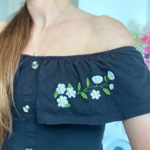 May include: Black off-the-shoulder top with a ruffled neckline and a floral embroidery design. The embroidery features white flowers and green leaves.