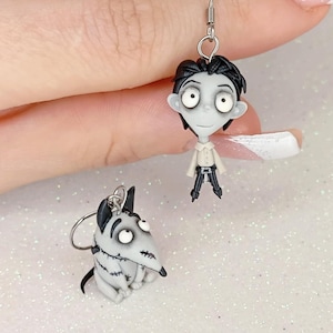 Puede incluir: Un par de pendientes hechos a mano con un personaje y su perro. El personaje tiene pelo negro, una camisa blanca y pantalones negros. El perro es gris y blanco con detalles negros. Los pendientes están en un gancho plateado.