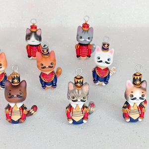 Op de afbeelding: Negen handgemaakte kattenfiguren verkleed als speelgoed soldaten. Elke kat draagt een rood en blauw uniform met gouden accenten en een ster op zijn hoed. De katten zijn allemaal in verschillende kleuren en hebben unieke gezichtsuitdrukkingen.