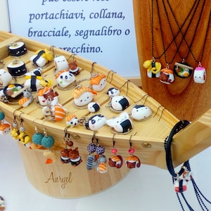 Puede incluir: Un expositor de madera con forma de barco lleno de amuletos hechos a mano con temática de sushi, incluyendo llaveros, collares y pendientes. Los amuletos están hechos de arcilla polimérica y presentan varios rollos de sushi, nigiri y otros alimentos japoneses. El expositor está decorado con el texto "Ogni ciondolo esposto può essere reso portachiavi, collana, bracciale, segnalibro o vecchino."