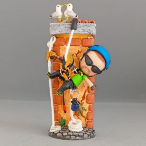 Puede incluir: Una figura caprichosa de un escalador descendiendo por una chimenea de ladrillo. El escalador lleva un casco azul, gafas de sol y equipo de escalada. Dos gaviotas se posan en la parte superior. Un pequeño lagarto y una bobina de cuerda están en la base.