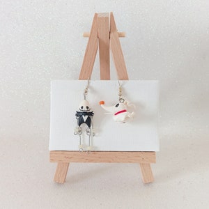 May include: A pair of dangle earrings featuring a black and white skeleton character and a white dog character. The earrings are displayed on a small wooden easel.
