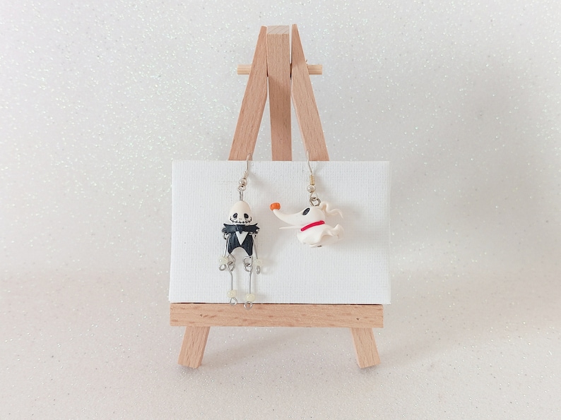 May include: A pair of dangle earrings featuring a black and white skeleton character and a white dog character. The earrings are displayed on a small wooden easel.