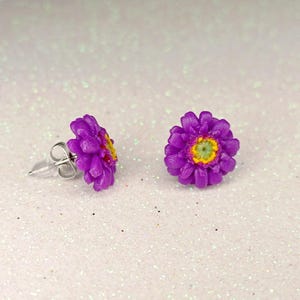 Può includere: Un paio di orecchini a bottone a forma di fiore viola con centri gialli e verdi. Gli orecchini sono realizzati in materiale stampato e hanno perni color argento. Gli orecchini sono su uno sfondo bianco glitterato.
