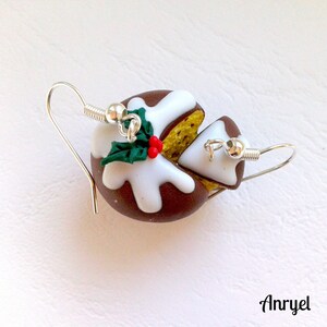 May include: A pair of earrings featuring a miniature Christmas pudding with a slice cut out, decorated with white icing and a green holly sprig with red berries. The earrings are made of polymer clay and have silver hooks.