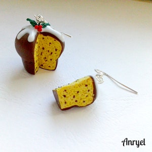 May include: A pair of earrings featuring a miniature panettone, a traditional Italian sweet bread, with a chocolate glaze and white icing. One earring is a whole panettone, while the other is a slice. The earrings are made of polymer clay and have silver hooks.