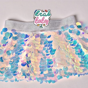 May include: A white iridescent sequin skirt with a silver elastic waistband. The skirt is made of small, round sequins that are arranged in a scale-like pattern. The sequins are iridescent, meaning they change color depending on the angle of light. The skirt is perfect for a mermaid costume or a fun, festive outfit.