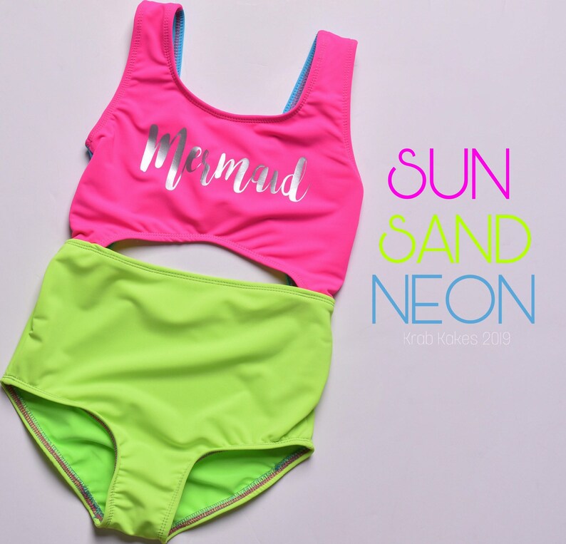 May include: A neon green and pink swimsuit with a cutout in the middle. The top of the swimsuit has a silver glitter "Mermaid" design. The text "SUN SAND NEON" is written in pink, yellow, and blue letters on the right side of the image. The text "Krab Kokes 2019" is written in blue letters below the text "NEON".