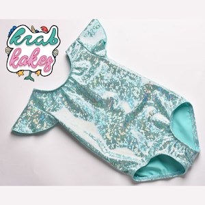 May include: A light blue leotard with iridescent sequins and short sleeves. The leotard has a scoop back and a turquoise lining. The image includes the text "krab kakes" in a colorful, cartoon-style logo.
